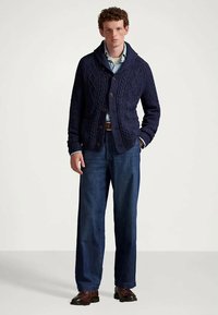 Mężczyzna w granatowym swetrze w warkocze noszącym jasnoniebieską koszulę, w szerokich dżinsach i brązowych skórzanych butach, stojący na tle jednolitym.