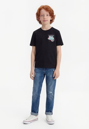 Dreng med rødt hår iført en sort t-shirt med grafik, blå opkørte jeans og hvide sneakers, stående foran en ensfarvet hvid baggrund.