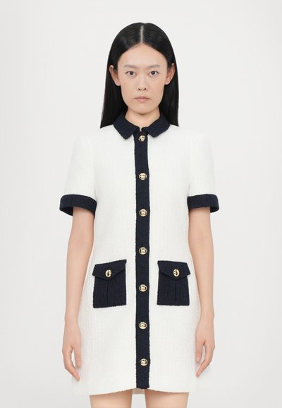 Abito bianco con colletto navy e tasche, caratterizzato da bottoni dorati. Tessuto testurizzato con maniche corte e una silhouette aderente.