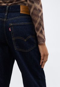 Personne portant un jean Levi's bleu foncé et une chemise à manches longues à carreaux marron, la main reposant près de la hanche sur un fond uni.
