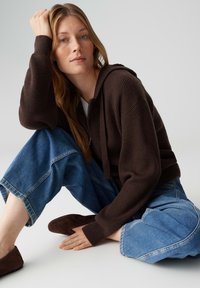 Hoodie marron côtelé avec cordons de serrage réglables, associé à un jean en denim bleu à jambes larges. Le modèle porte des chaussures marron en daim à enfiler.