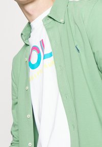 Mężczyzna ubrany w jasnozieloną koszulę zapinaną na guziki, noszącą białą koszulkę pod spodem, z kolorowym napisem "Polo Ralph Lauren".