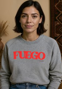 Grijze sweatshirt met "FUEGO" in vet rood lettertype. Heeft een ronde halslijn en ribgebreide manchetten. De achtergrond bevat een patroontextiel en een plant.