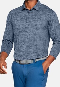 Mann trägt ein blaues, strukturiertes Langarm-Polohemd, blaue Hose und einen geflochtenen grauen Gürtel, mit einer Hand in der Tasche.