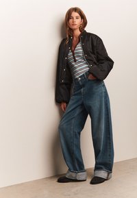 Zwarte bomberjack, gestreept shirt met lange mouwen, wijd uitlopende blauwe spijkerbroek met omgeslagen zoom en zwarte schoenen. Soepele texturen en casual design.