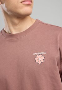 Ruskea puuvillapaita pyöreällä kaula-aukolla, jossa on vaaleanpunainen kukkakuva ja valkoinen "CALIFORNIA" -teksti sen yläpuolella.