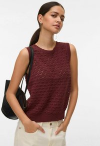 Femme aux cheveux foncés attachés en queue de cheval, portant un haut en maille sans manches marron rouge, un pantalon crème, un sac à main noir et des boucles d'oreilles argentées, les mains dans les poches.