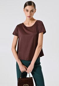 Braune Kurzarmbluse mit rundem Ausschnitt, aus glattem Stoff. Kombiniert mit dunkelgrünen Hosen und einer strukturierten braunen Handtasche.