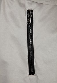 Bolsillo con cremallera en tela gris. La cremallera es negra con un tirador metálico y dientes. La tela tiene una textura suave con un ligero brillo.