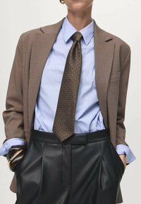 Bruin geruite blazer gedragen over een lichtblauw gestreept shirt, gecombineerd met een bruine gemusterde stropdas en hoogwaisted zwarte leren broek.