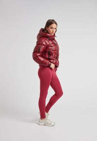 Giacca a vento rossa con cappuccio, dotata di chiusura con zip e tasche laterali. Abbinata a leggins rossi scuri testurizzati e scarpe da ginnastica argentate.