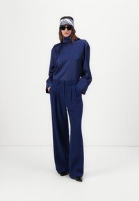 Top in satin navy a maniche lunghe con colletto alto, abbinato a pantaloni a gamba larga con pences. Scarpe nere a punta e fascia per capelli fantasia.