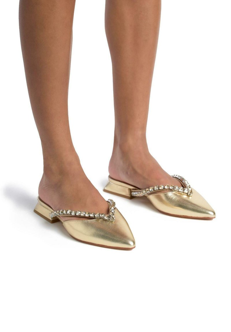 Des mules dorées métalliques à enfiler avec un bout pointu et une sangle décorative agrémentée de strass traversant le cou-de-pied. Design à semelle plate.