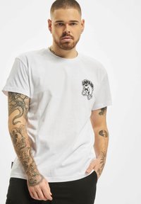 Camiseta de algodón blanca con un gráfico negro del logo "UNFAIR" en el lado izquierdo del pecho, mangas cortas y un corte relajado. Color sólido, diseño minimalista.