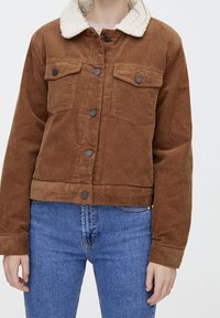 Persona che indossa una giacca di velluto a coste marrone con un colletto in pile bianco e jeans di denim blu, in piedi contro uno sfondo semplice.