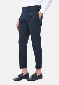Pantaloni a pieghe blu navy con una cintura ampia e due bottoni. Presentano tasche laterali e orli accorciati. Abbinati a mocassini scuri.