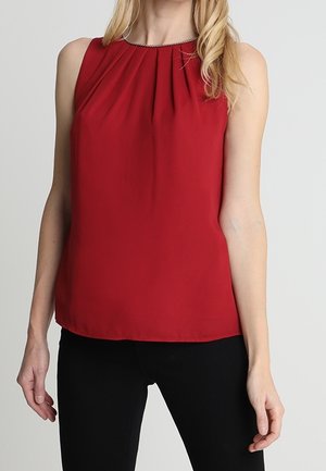 Mouwloze rode blouse met plooien bij de hals, gedragen met zwarte broek bij een staand persoon tegen een effen achtergrond.