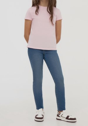 Dziewczyna stoi z rękami za plecami, ubrana w jasnoróżową koszulkę z krótkim rękawem, niebieskie dżinsy i białe trampki z brązowymi detalami.