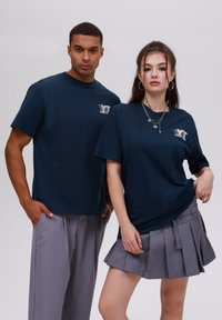 Tumši zili īsām piedurknēm t-krekli ar baltu logotipu. Sieviešu modelis ģērbies pelēkā svārku plānojumā, vīriešu modelis pelēkās biksēs. Vienkāršs dizains.