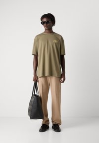 Oliven grøn oversized t-shirt med gult logo, beige bukser, sorte solbriller og en tekstureret sort tote bag. Skoene er mørke og kraftige.