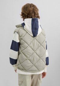 Niño con pelo rizado que lleva un chaleco acolchado verde sobre una sudadera blanca con rayas anchas azules, de pie de espaldas sobre un fondo gris.