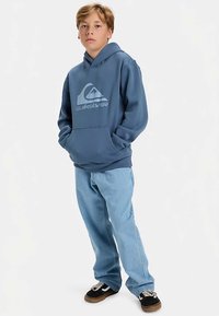 Boy standing with hands in pockets wearing a blue Quiksilver hoodie, light blue jeans, and black sneakers on a white background.