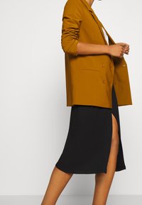 Senapsgul dubbelknäppt kavaj med slag, parat med en svart midiklänning med sidofickor, som framhäver släta texturer.