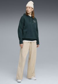 Felpa verde scuro con tasca frontale e cordini, abbinata a pantaloni larghi color beige chiaro e sneakers bianche, completata da un berretto di colore crema.