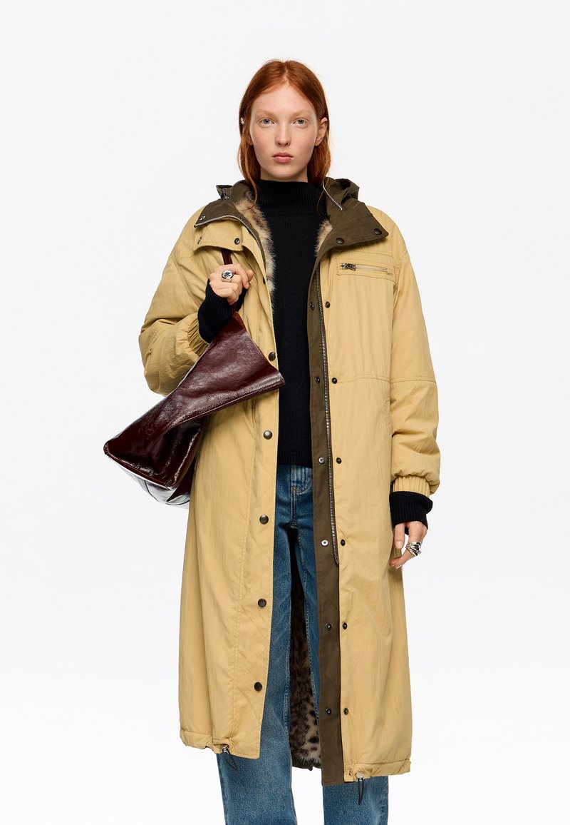 Mujer joven con cabello rojo que lleva un abrigo largo beige sobre un suéter negro y vaqueros azules, sosteniendo un bolso de cuero rojo oscuro.