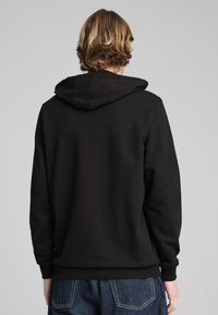 Zwarte hoodie met ribgebreide manchetten en onderrand. Zachte textuur, eenvoudige ontwerp. Model met de rug naar de camera, toont de details van de achterkant en de capuchon.
