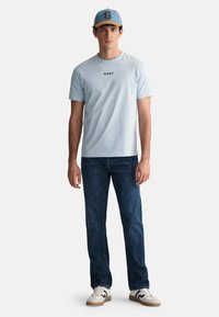 Tricou din bumbac de culoare albastru deschis cu logo-ul "GANT", asortat cu blugi de culoare albastru închis. Purtați cu o șapcă de culoare albastru deschis, având o vizieră galbenă și litera "G".