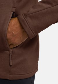 Brauner Fleece-Pullover mit einer seitlichen Reißverschlusstasche. Verfügt über gerippte Bündchen und einen geraden Saum. Sichtbare glatte, weiche Textur.