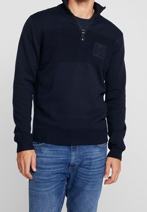 Mężczyzna ubrany w granatowy sweter z półzamkiem i teksturowanym górnym panelem oraz jasnoniebieskie dżinsy, na jednolitym jasnoszarym tle.