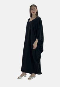Czarna maxi sukienka z dekoltem w kształcie litery V i rękawami nietoperzowymi. Wykonana z miękkiego materiału, z falbankami i luźnym krojem. Model nosi białe sandały na obcasie.