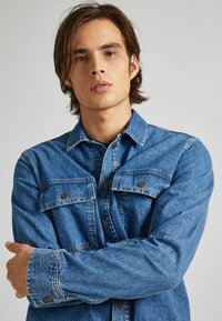 Modra jeans jakna z dvema žepoma na prsih, gumbnimi zapiranji in vidnimi šivi. Ima klasičen ovratnik in sproščen kroj.