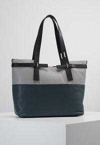 Borsa tote in pelle grigio e blu scuro a due toni con spallacci neri regolabili, esposta su un pedestal bianco.