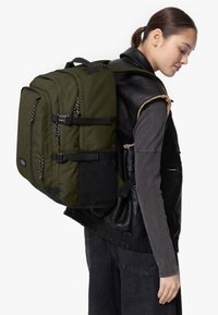 Olivgrön ryggsäck med svarta detaljer, flera fack, justerbara remmar och en strukturerad yta, bärs på en svart och grå outfit.