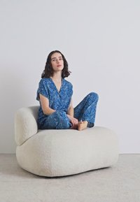 Iškirpta balta boucle kėdė su minkšta, tekstūruota paviršiumi. Žmogus mėlynais raštuotais laisvalaikio drabužiais sėdi atsipalaidavęs, sukryžiavęs kojas, ant kėdės.