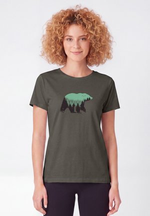 Frau mit lockigem Haar trägt ein dunkelgrünes T-Shirt mit einer Bärensilhouette, die mit Bäumen und einer Berglandschaft gefüllt ist.