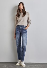 Suéter beige claro, jeans rectos azules con textura deslavada y zapatillas blancas. Conjunto casual con un diseño minimalista y acentos simples.