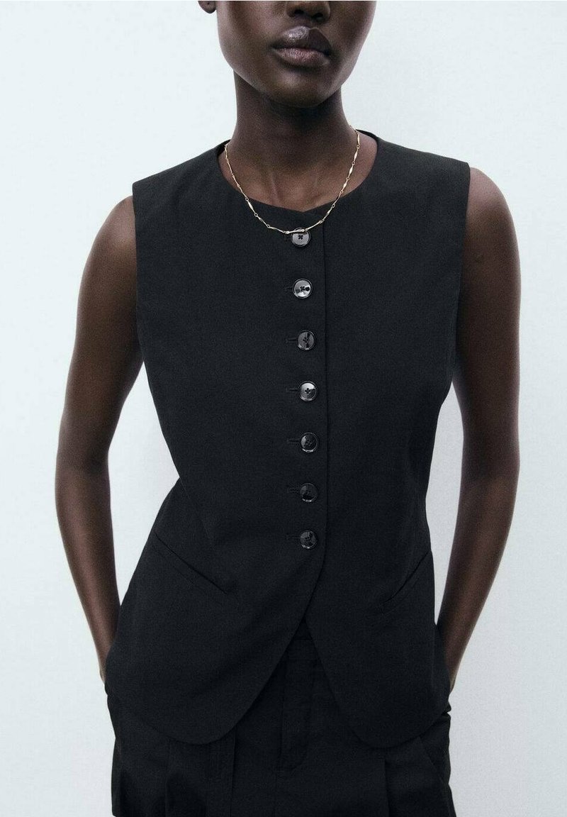 Personne portant un gilet noir sans manches boutonné avec des poches et un délicat collier en chaîne dorée, debout devant un fond uni.