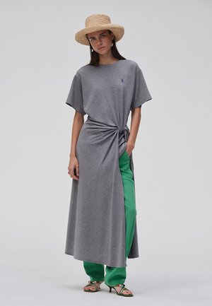 Kvinne iført en grå knyttet kjole over grønne bukser, grønne høyhælte sandaler og en stråhatt, stående mot en ensfarget lys bakgrunn.