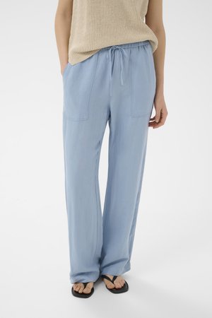 Henkilö pukeutunut vaaleansiniset leveälahkeiset housut, joissa nyörivyötärö, beige hihaton neulepaita ja mustat varvassandaalit valkoisella taustalla.