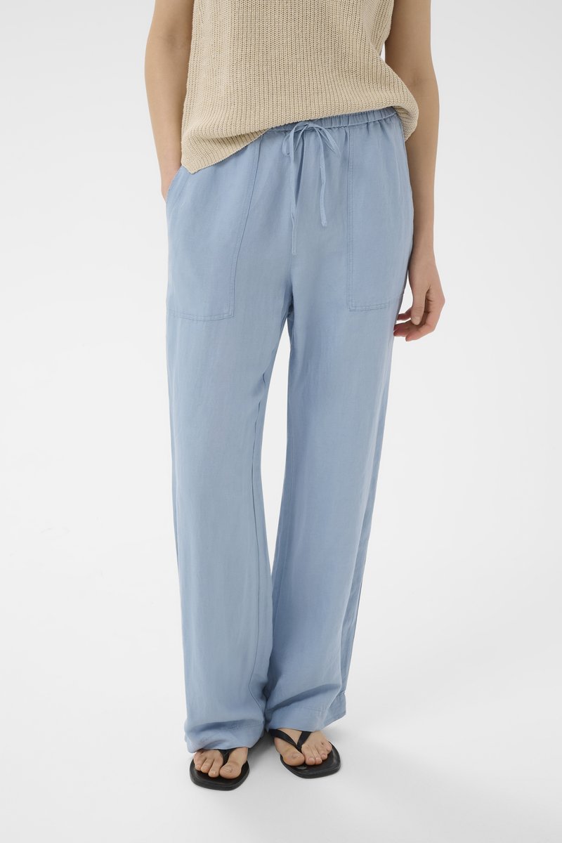 Persona che indossa pantaloni azzurri a gamba larga con coulisse in vita, top beige senza maniche in maglia e sandali infradito neri su sfondo bianco.