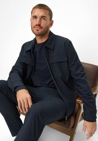 Marine, met rits, jacket met twee borstzakken, getailleerde pasvorm en gladde stof. Gecombineerd met een bijpassend marineblauw shirt en broek.