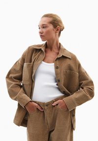 Giacca di corduroy marrone con righe verticali ben visibili, chiusura a bottoni e tasca sul petto. Abbinata a pantaloni di corduroy coordinati e a una canotta bianca.