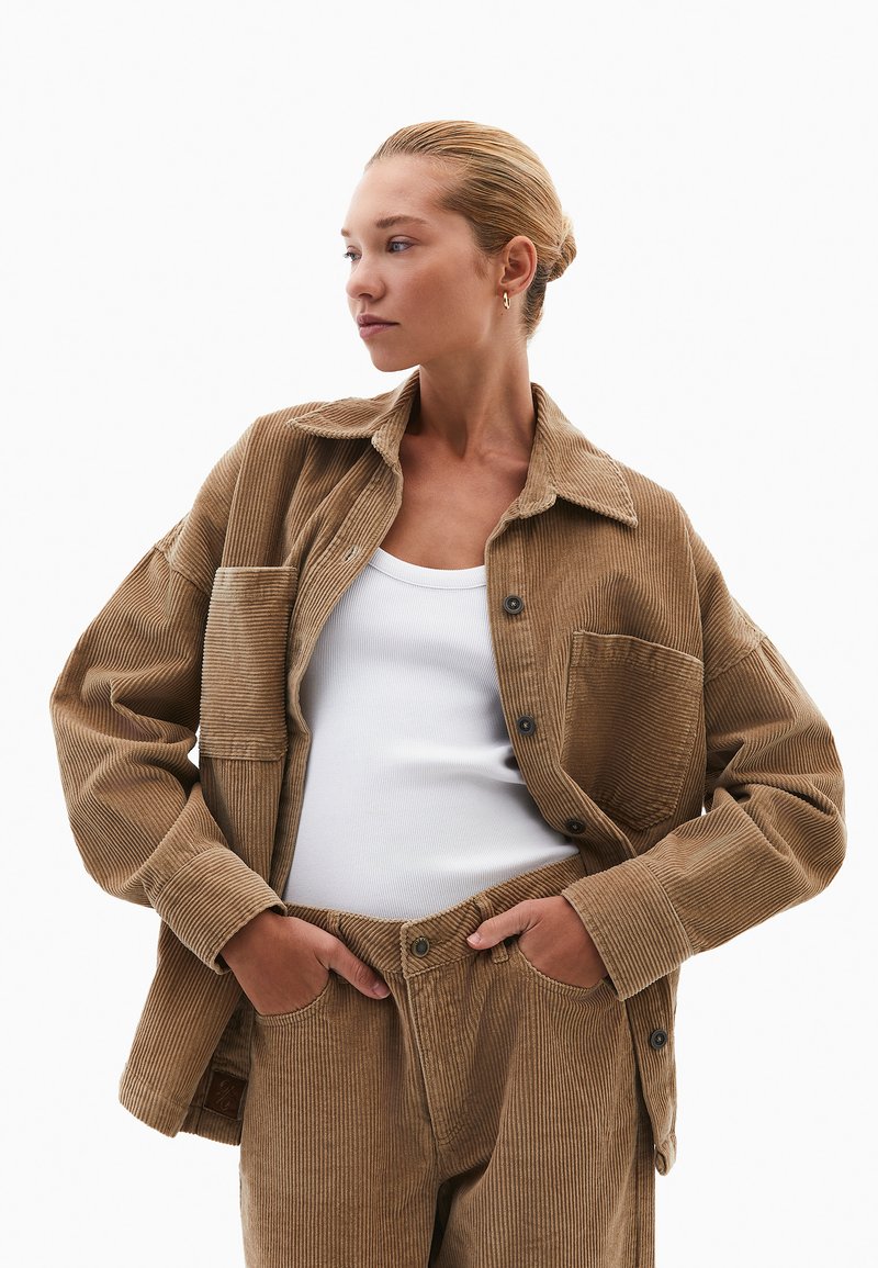 Giacca di corduroy marrone con righe verticali ben visibili, chiusura a bottoni e tasca sul petto. Abbinata a pantaloni di corduroy coordinati e a una canotta bianca.