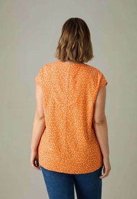 Blusa arancione con stampa floreale, maniche corte a cappuccio e orlo arrotondato. Presenta una cucitura centrale sul retro, realizzata in tessuto leggero.