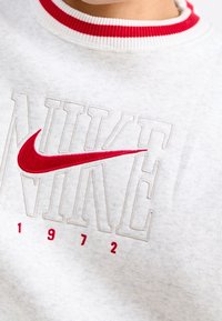 Felpa grigia con colletto a righe rosse e bianche, con logo "NIKE" ricamato in bianco e rosso e l'anno "1972" in rosso sotto.