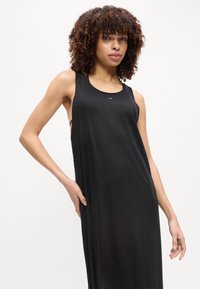 Γυναίκα με σγουρά μαλλιά, ντυμένη με μαύρο φόρεμα χωρίς μανίκια, στέκεται με το ένα χέρι στη μέση της μπροστά σε απλό λευκό φόντο.